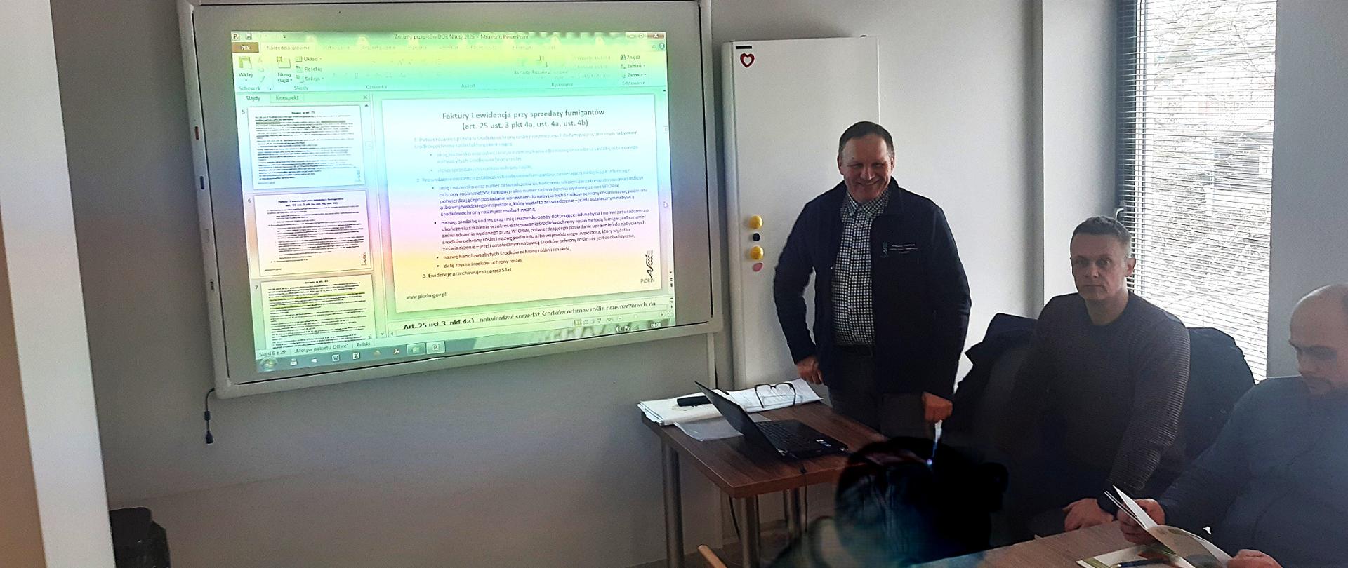 Uczestnicy szkolenia siedzą na sali konferencyjnej, wykładowca stoi przodem do słuchaczy i prowadzi wykład