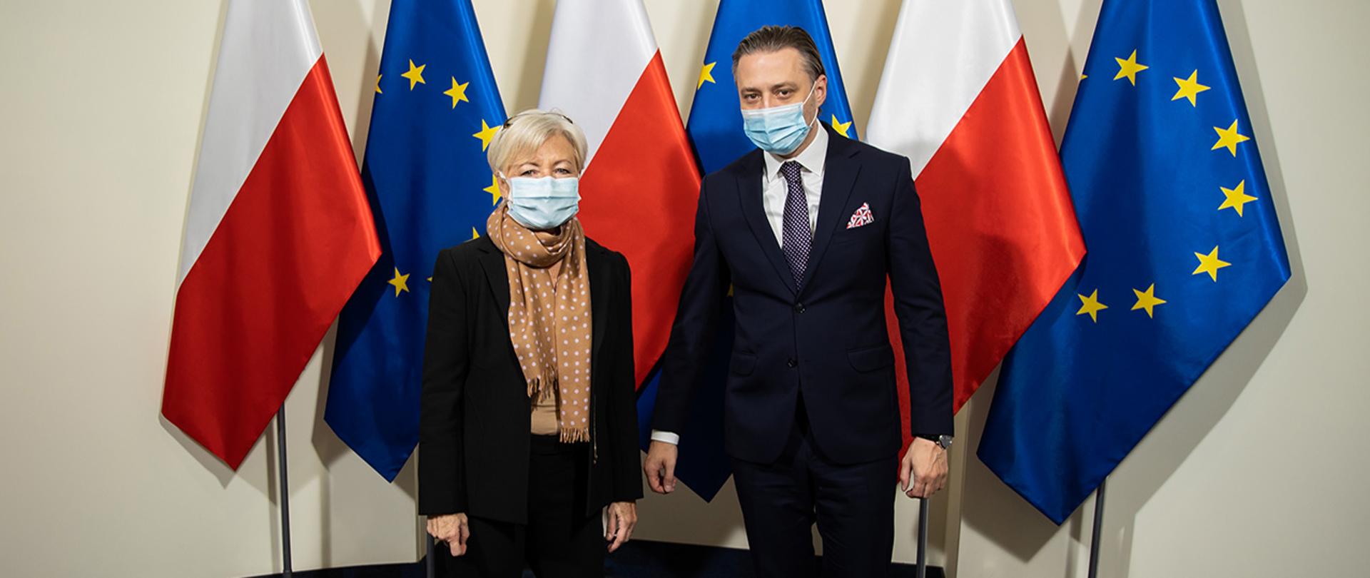 Na zdjęciu widać wiceministra Bartosza Grodeckiego z Monique Pariat Dyrektor Generalną w Dyrekcji Generalnej ds. Migracji i Spraw Wewnętrznych Komisji Europejskiej stojących na tle flag Polski i UE.