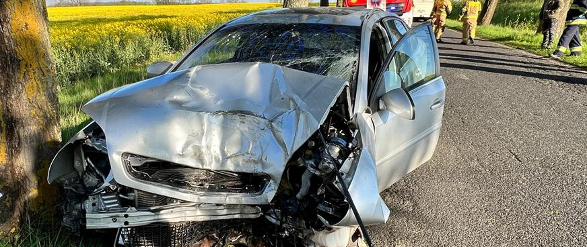 Zdjęcie przedstawia srebrny samochód po uderzeniu w drzewo stojący na poboczu drogi.