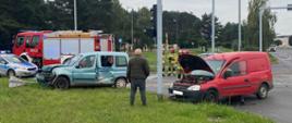 Dwa samochody osobowe z uszkodzonymi przodami stoją jeden na jezdni drugi na poboczu. W tle samochody strażacki oraz strażacy.