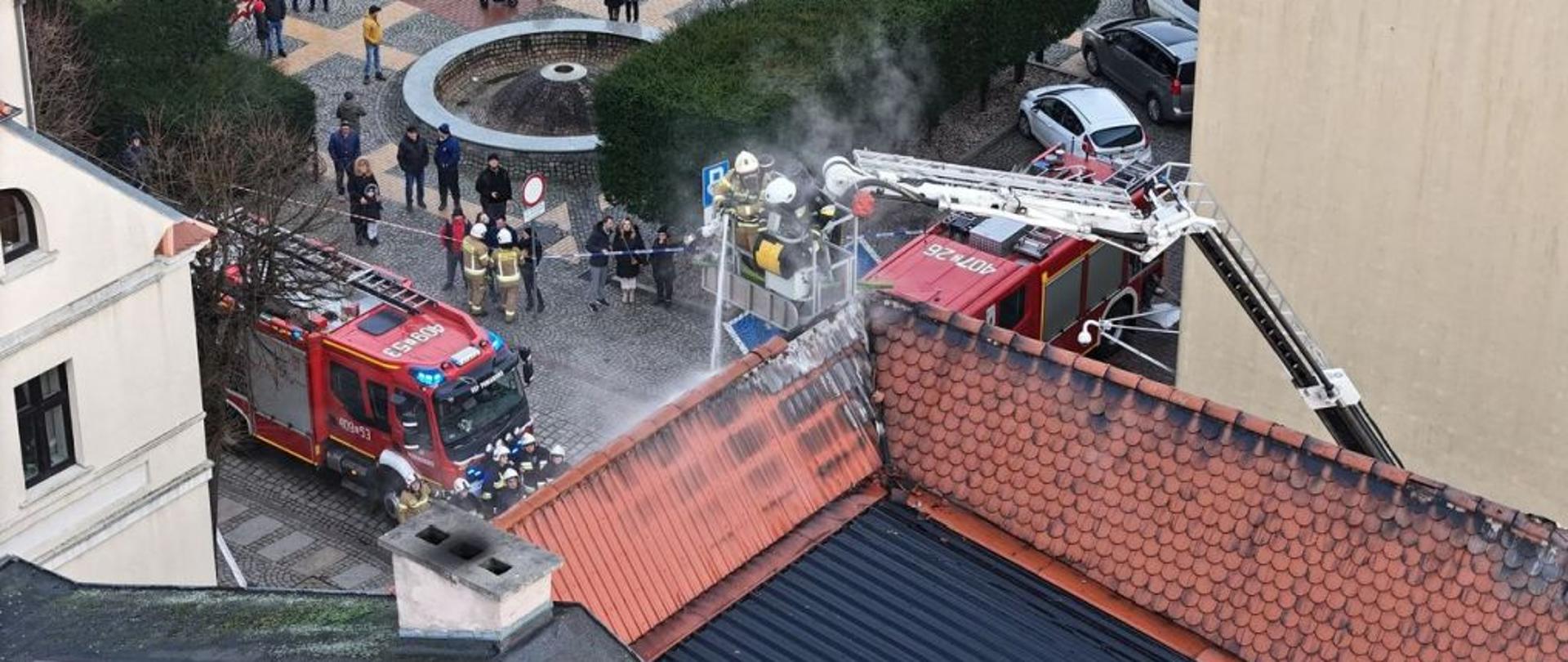 Zdjęcie wykonane z drona. Centralny punkt zdjęcia stanowi kosz podnośnika i strażacy gaszący pożar.