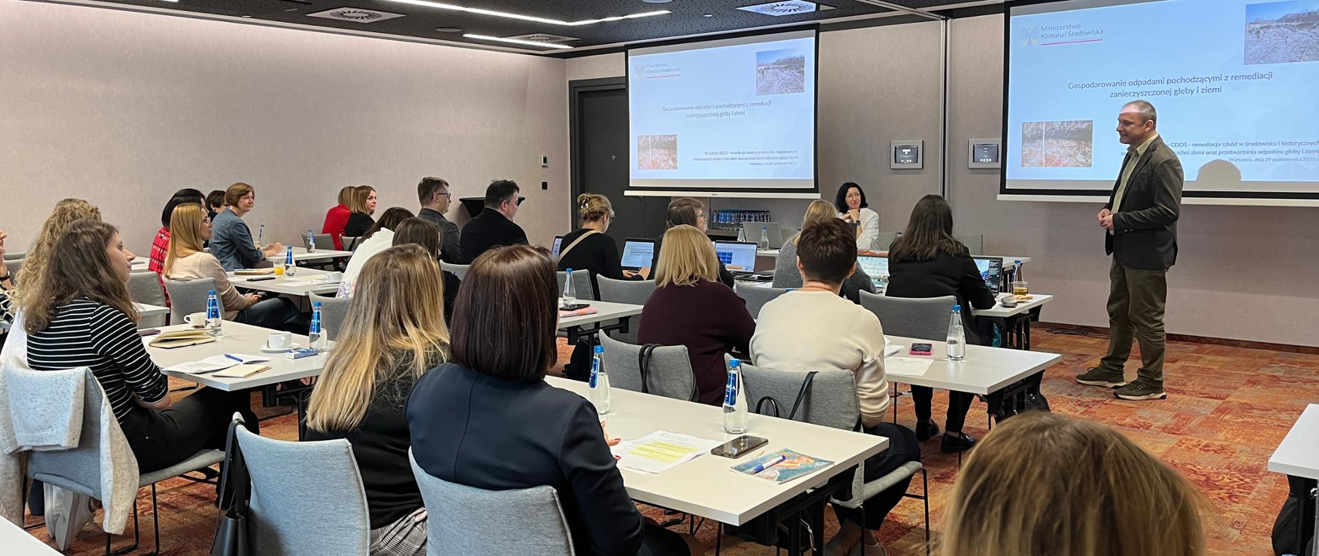 Widok sali konferencyjnej. Na pierwszym planie widoczni są uczestnicy warsztatów, którzy siedzą plecami do obiektywu. Prelegent i ekran jest w dalszej części kadru. Prelegent (mężczyzna w ciemnej marynarce i spodniach w kolorze ciemnej zieleni) stoi po prawej stronie kadru. Uczestnicy siedzą w rzędach przy białych stołach. Skupieni są na prezentacji. Kilka osób ma przed sobą laptopy i robi notatki.