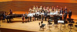 Chór PSM i Chór Miejski stojący na scenie sali koncertowej PSM oraz po lewej stronie akompaniator przy fortepianie i Pani Dyrygent