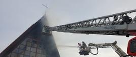 Widok kościoła od frontu. Strażacy polewają wodę z drabiny mechanicznej i podnośnika hydraulicznego na część dachu, nie widać już płomieni, tylko dym i rozbite witraże.