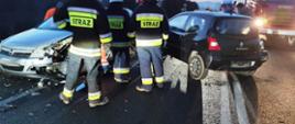 Dwa rozbite samochody stojące na drodze a pomiędzy nimi srtoi trzech strażaków.