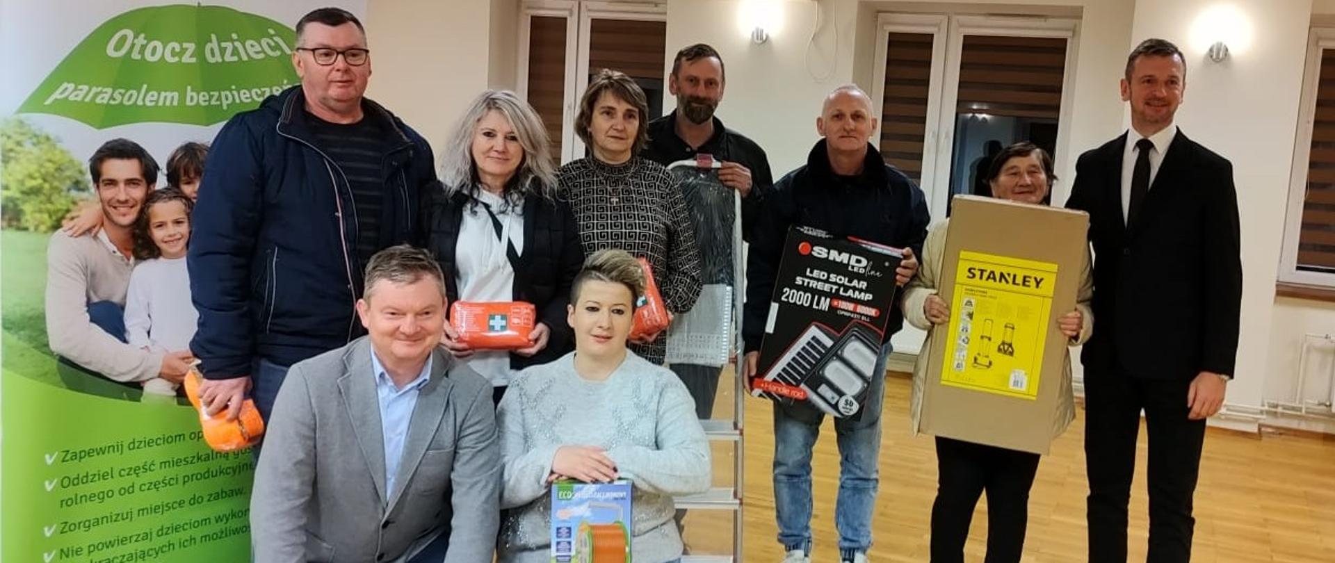 W sali na tle rollupu KRUS stoją trzy kobiety i czterech mężczyzn. Sześcioro z nich trzyma w rękach nagrody. Przed nimi przykucnęli mężczyzna i kobieta. Kobieta także trzyma nagrodę.