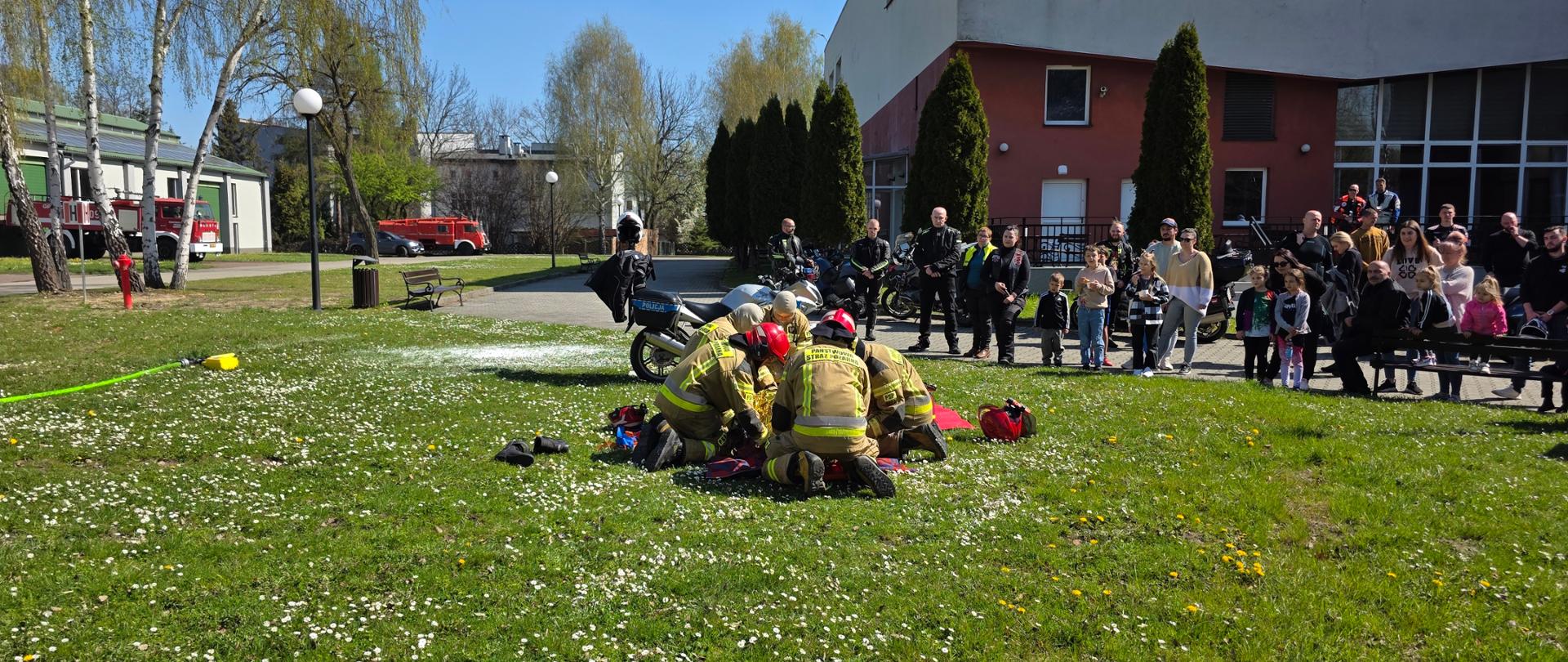 To zdjęcie dokumentuje kulminacyjny moment pokazu na terenie **Centralnego Muzeum Pożarnictwa**, ukazując to, co w tym wydarzeniu było najważniejsze – bezpośrednią interakcję służb z publicznością.
Oto szczegółowy opis tej perspektywy:
* **Akcja ratunkowa w centrum uwagi:** Grupa strażaków w piaskowych mundurach bojowych jest w trakcie wykonywania precyzyjnych czynności ratunkowych przy poszkodowanym. Wokół nich na trawie widać rozłożony sprzęt, w tym jaskrawoczerwoną torbę medyczną i kask, co dodaje scenie realizmu.
* **Publiczność:** To ujęcie najlepiej pokazuje frekwencję podczas akcji „Bezpieczny start”. Po prawej stronie, na placu przed czerwonym budynkiem muzeum, zgromadziła się liczna grupa obserwatorów. Są wśród nich zarówno dorośli motocykliści w skórzanych strojach, jak i rodziny z dziećmi. Wszyscy z zaciekawieniem śledzą każdy ruch ratowników.
* **Elementy edukacyjne w tle:** * Po lewej stronie kadru widoczny jest rozciągnięty na trawie zielony wąż strażacki (linia gaśnicza) oraz charakterystyczny czerwony hydrant.
* W tle, przy alejkach, parkują kolejne motocykle uczestników inauguracji sezonu.
* **Architektura i otoczenie:** Zdjęcie obejmuje fragment nowoczesnego, czerwono-szarego budynku muzeum oraz rzędy wysokich tuj, które oddzielają strefę pokazową od parkingu. Wysokie brzozy z młodymi liśćmi i pełne słońce tworzą przyjazną, wiosenną atmosferę pikniku edukacyjnego.
To ujęcie doskonale podsumowuje cel wydarzenia – edukację poprzez pokaz, który przyciągnął lokalną społeczność i fanów jednośladów, ucząc ich, jak cenne mogą być pierwsze minuty po wypadku.