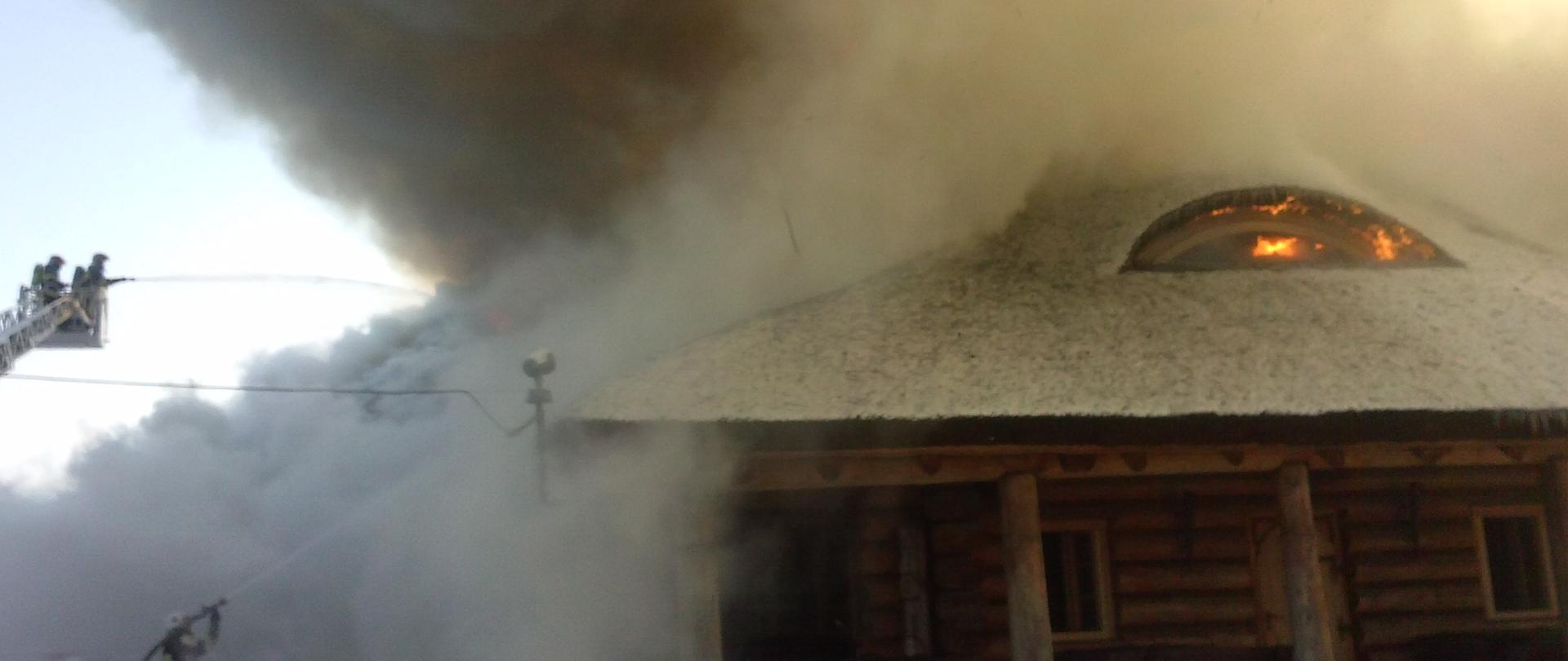 Zdjęcie przedstawia pożar budynku. Na zdjęciu widoczni są strażacy gaszący pożar. 