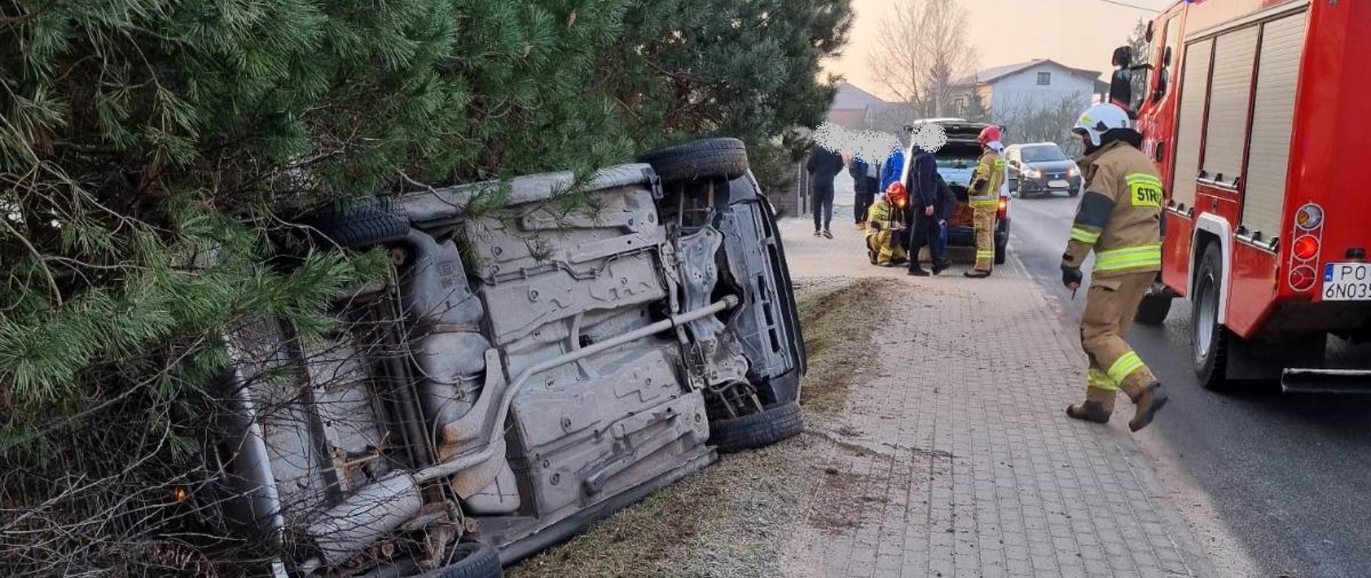Zdjęcie przedstawia samochód osobowy leżący na boku w przydrożnym rowie, oparty o ogrodzenie posesji. Na zdjęciu widać również samochód strażacki oraz strażaków. Samochód oparty o ogrodzenie przy którym znajdują się drzewa. 