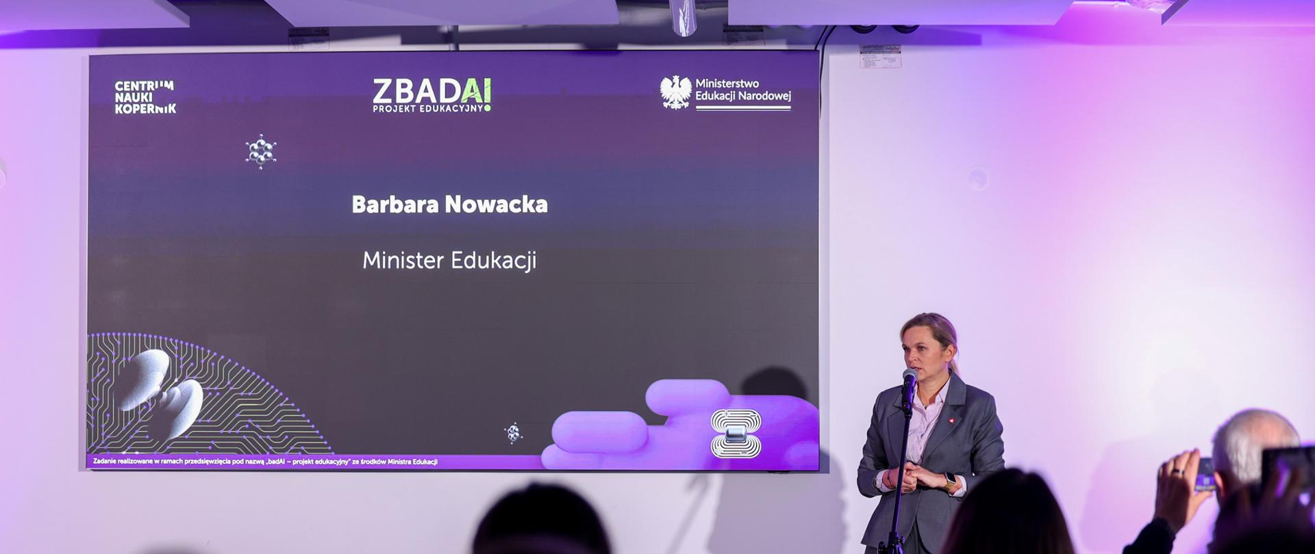 Zdjęcie przedstawia Minister Edukacji Barbarę Nowacką, mówiącą do ludzi siedzących przed nią. Za Minister Edukacji znajduje się duży ekran, na którym wyświetlana jest prezentacja. 