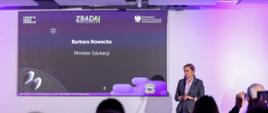 Zdjęcie przedstawia Minister Edukacji Barbarę Nowacką mówiącą do ludzi siedzących przed nią. Za Minister Edukacji znajduje się duży ekran, na którym wyświetlana jest prezentacja.