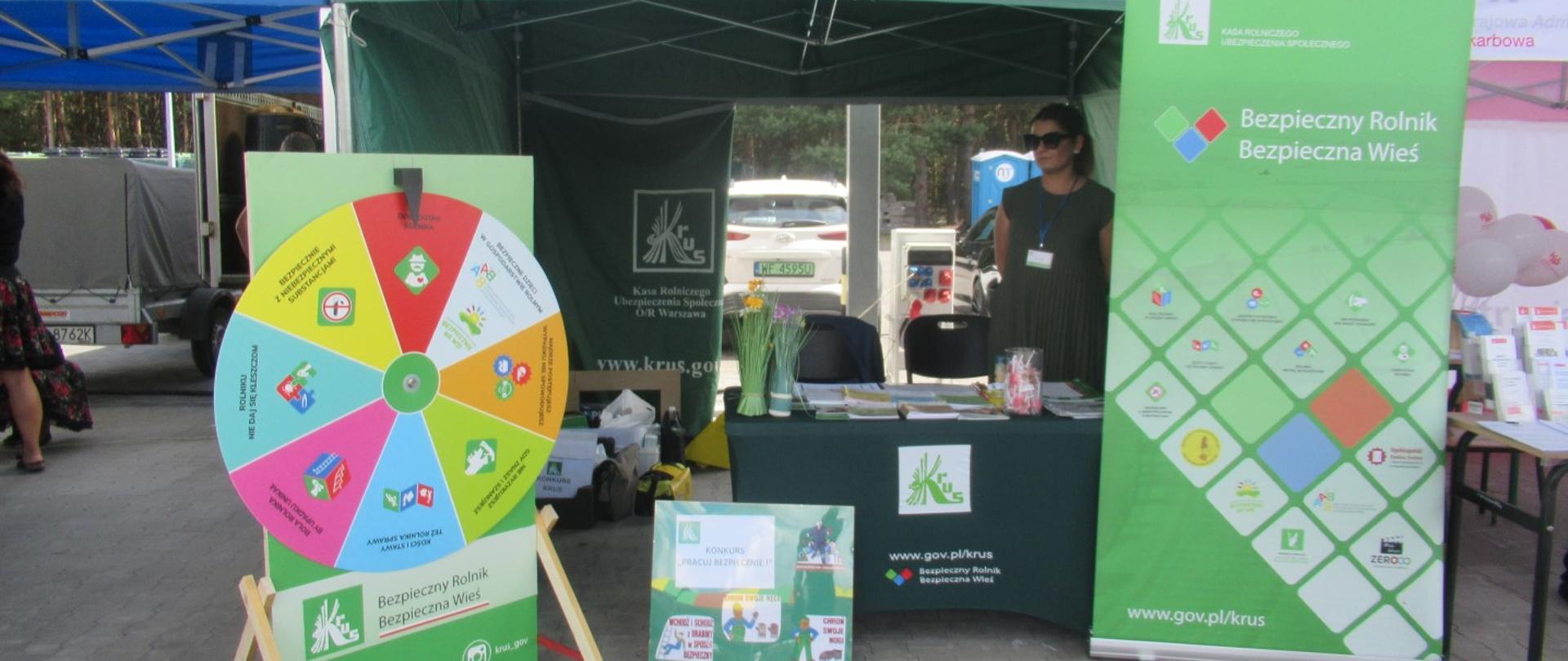 W namiocie stół nakryty zielonym obrusem z logo KRUS. Na stole broszury i ulotki informacyjne. Po prawej stronie namiotu baner z napisem „bezpieczny rolnik bezpieczna wieś”. Po lewej stronie namiotu koło bezpieczeństwa. W namiocie kobieta ubrana w zieloną sukienkę.