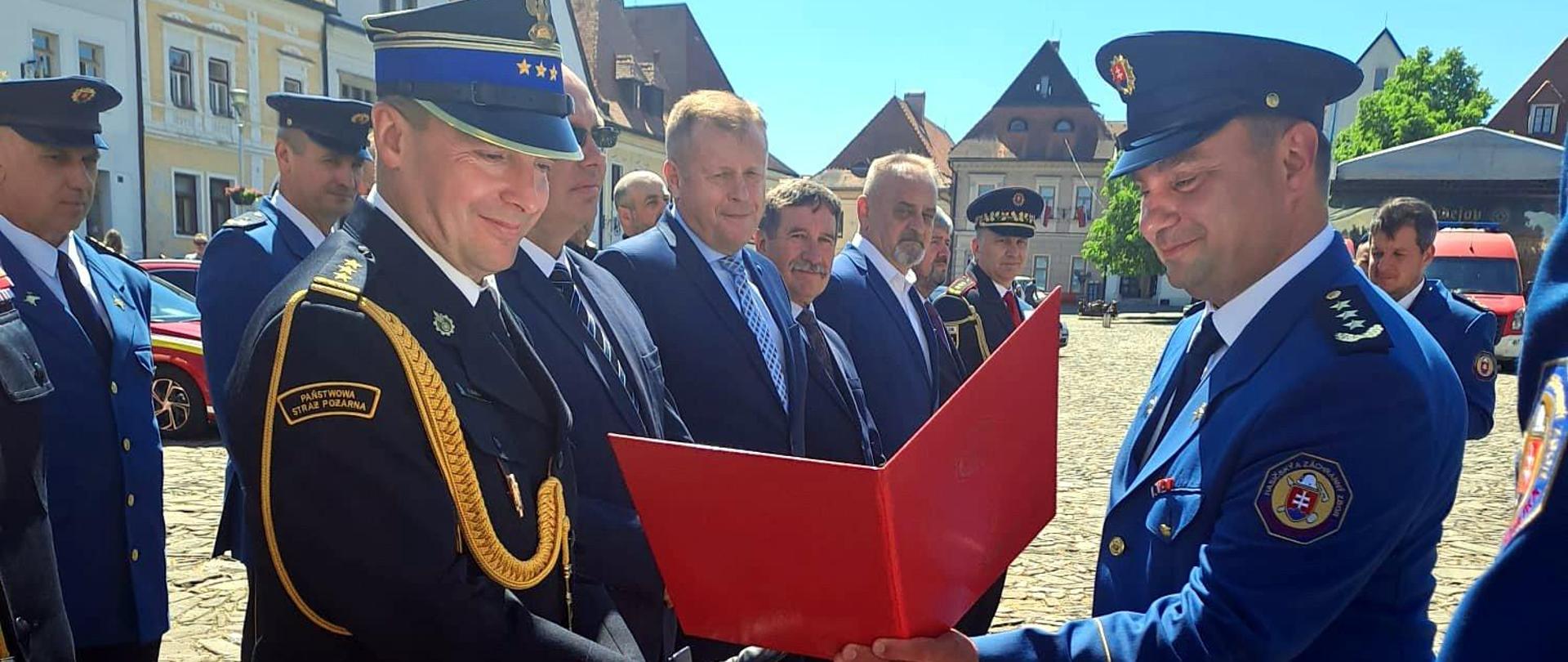 Zdjęcie zrobione na zewnątrz, na miejskim rynku podczas uroczystych obchodów dnia strażaka. Na zdjęciu stoją zaproszeni gości, uczestnicy uroczystości. Wśród nich słowaccy strażacy i oficerowie Państwowej Straży Pożarnej.