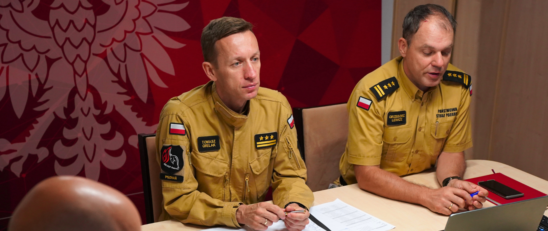 Na zdjęciu Pan Komendant i Naczelnicy wydziałów siedzą przy stole, przed telewizorem, podczas wideokonferencji, w pomieszczeniu, W tle znajduje się ścianka z napisem Państwowa Straż Pożarna.