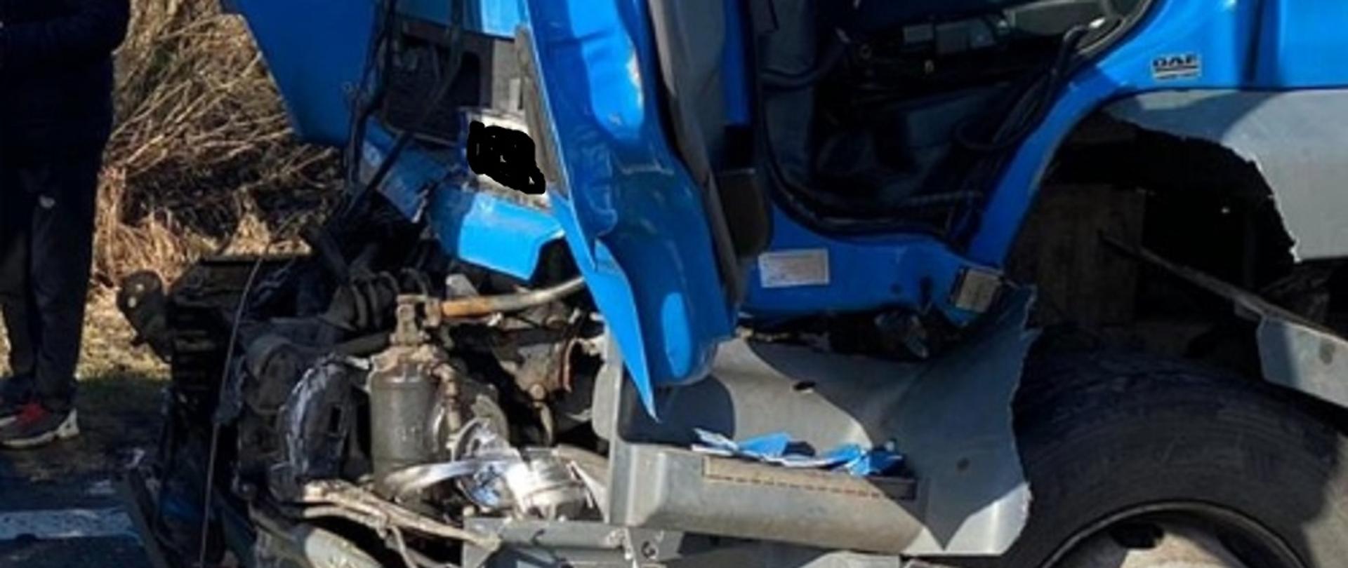 Samochód ciężarowy niebieski z uszkodzonym przodem kabiny pod wpływem zdarzenia.