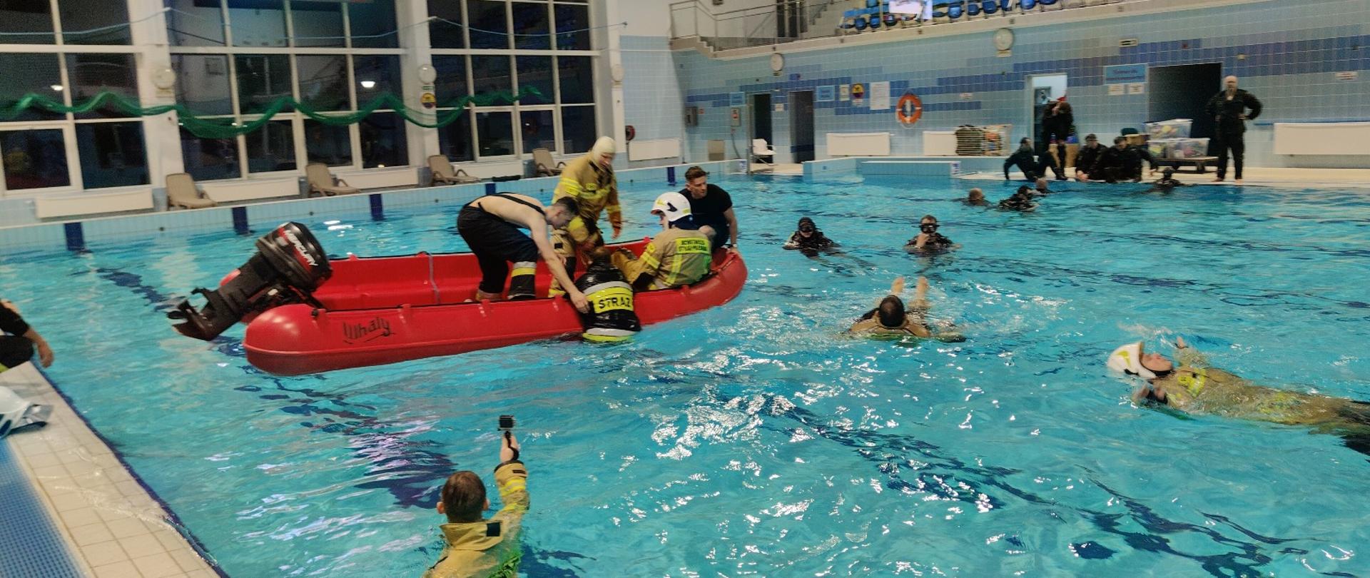 Strażacy pływają łodzią po basenie i w ramach założenia podejmują strażaków z wody. 