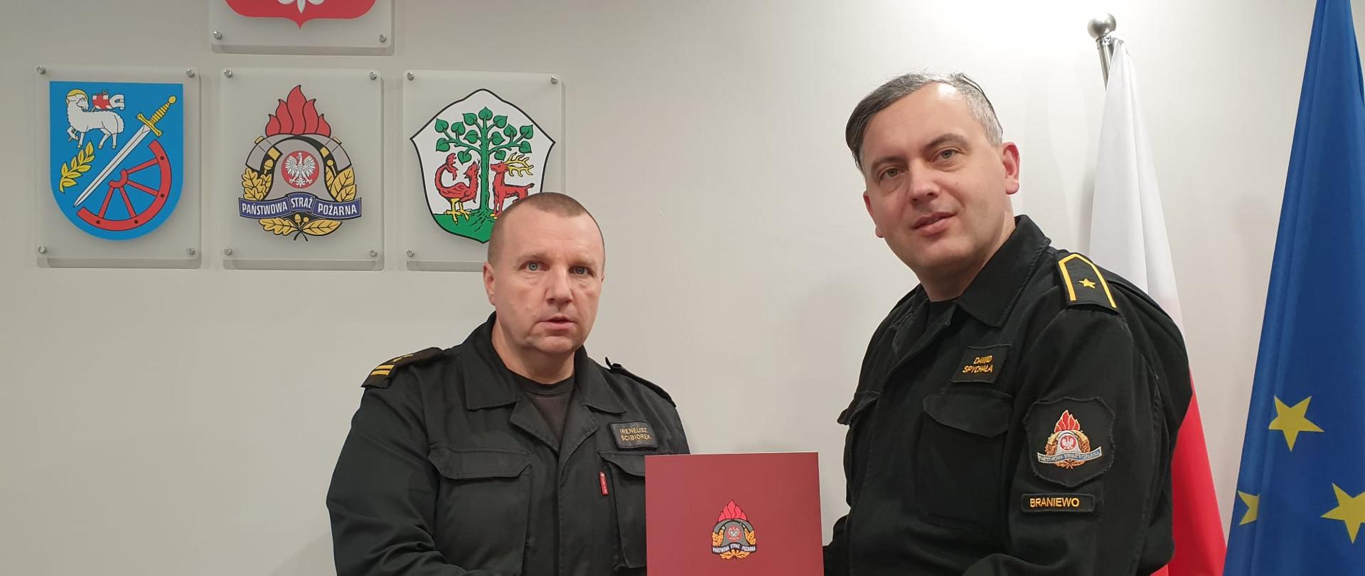 Pomieszczenie biurowe, strażacy w mundurach czarnych. Czerwona teczka w rękach. Z prawej strony Flagi, Polska i UE.