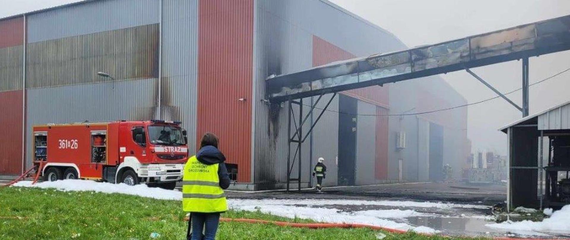 Na pierwszym planie Inspektor Ochrony Środowiska, na drugim planie samochód strażacki i strażak na tle spalonej hali