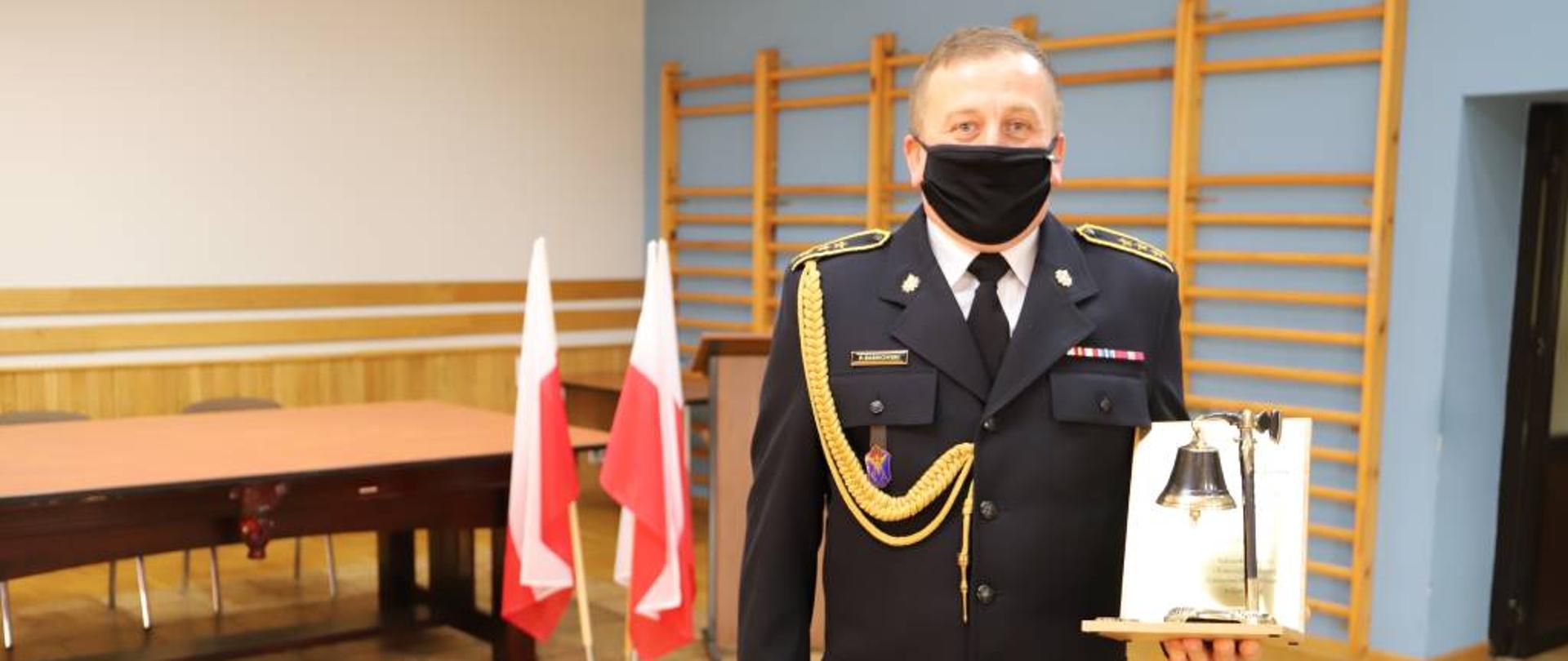 Zdjęcia przedstawia aspiranta sztabowego Pawła Mariusza Sasinowskiego w mundurze wyjściowym ze sznurem galowym trzymające w lewej ręce pamiątkowy dzwon, a w prawej kwiaty.