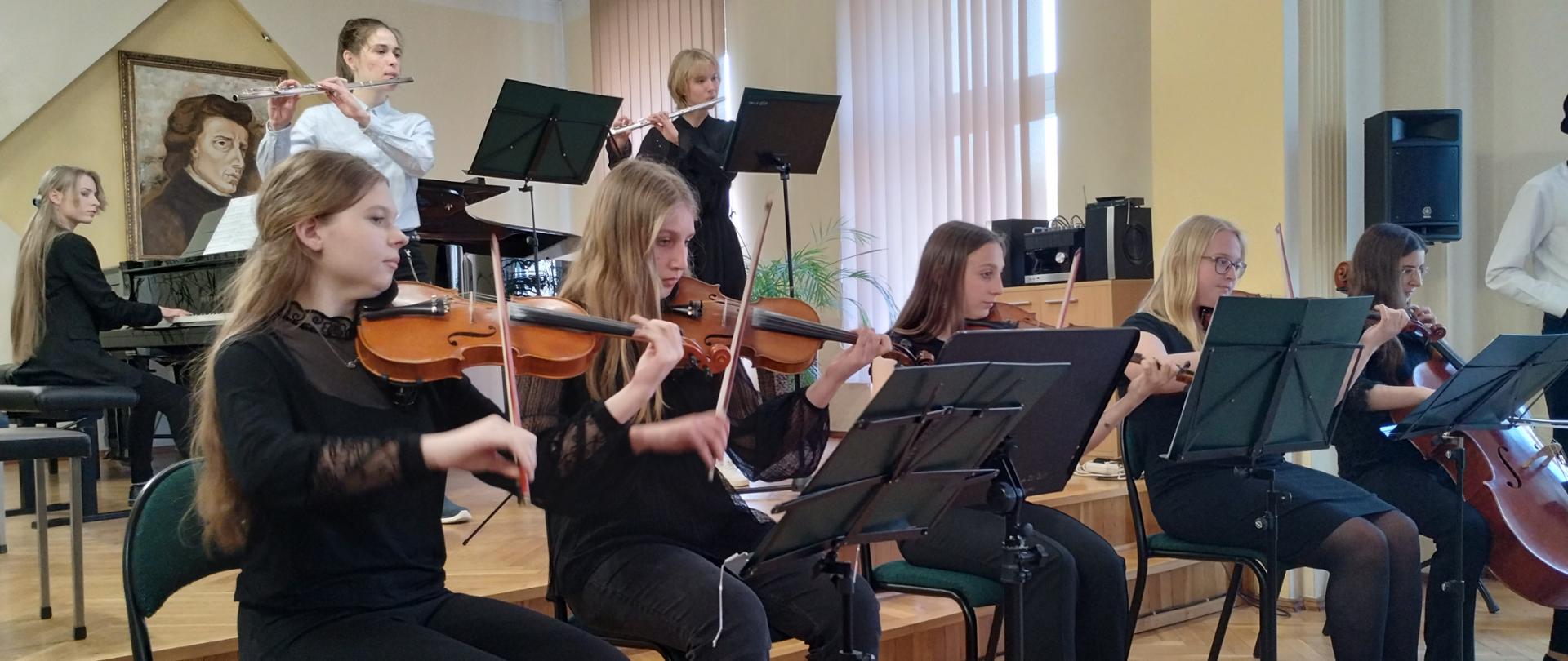 Zdjęcie przedstawia grupę uczennic 4 z nich grająca na skrzypcach i jedna na wiolonczeli w rzędzie, dwie na fletach poprzecznych i jedna na fortepianie podczas majowego koncertu w Kłodzku