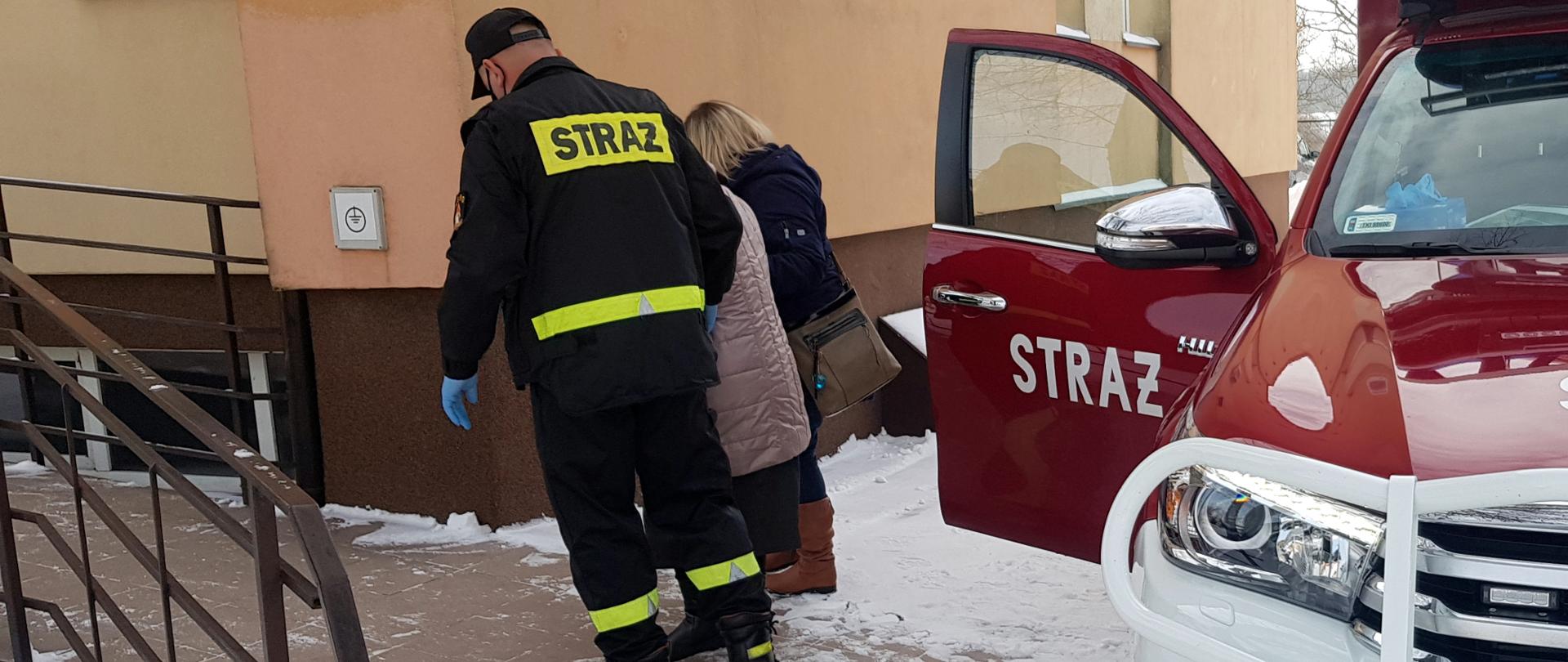 Na zdjęciu znajduje się samochód strażacki z otwartymi drzwiami a obok niego strażak prowadzący starszą osobę do punktu szczepień.