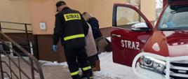 Na zdjęciu znajduje się samochód strażacki z otwartymi drzwiami, a obok niego strażak prowadzący starszą osobę do punktu szczepień.