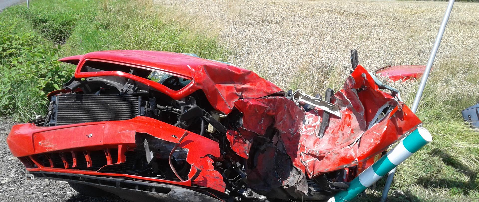 Zniszczony czerwony samochód osobowy po zderzeniu z innymi pojazdami. Pojazd na poboczu drogi, częściowo w rowie. W tle łan zboża.