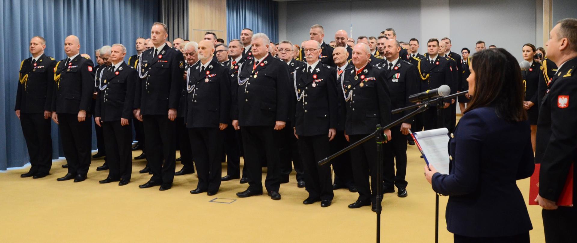 Uroczysta zbiórka z okazji wręczenia odznaczeń i nadania wyższych stopni. Na zdjęciu strażacy stojący w rzędach przed wręczeniem odznaczeń i nadaniem wyższych stopni.