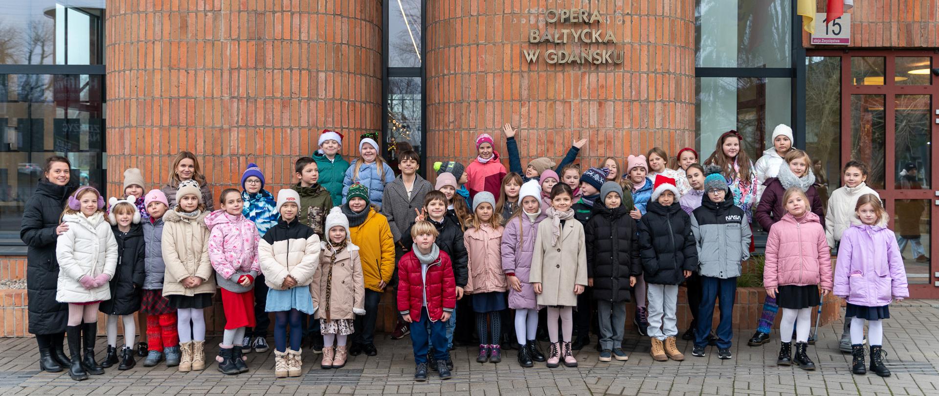 Przed budynkiem Opery Bałtyckiej pozuje liczna grupa uczniów wraz z opiekunami. Wszyscy stoją ubrani w kolorowe kurtki.