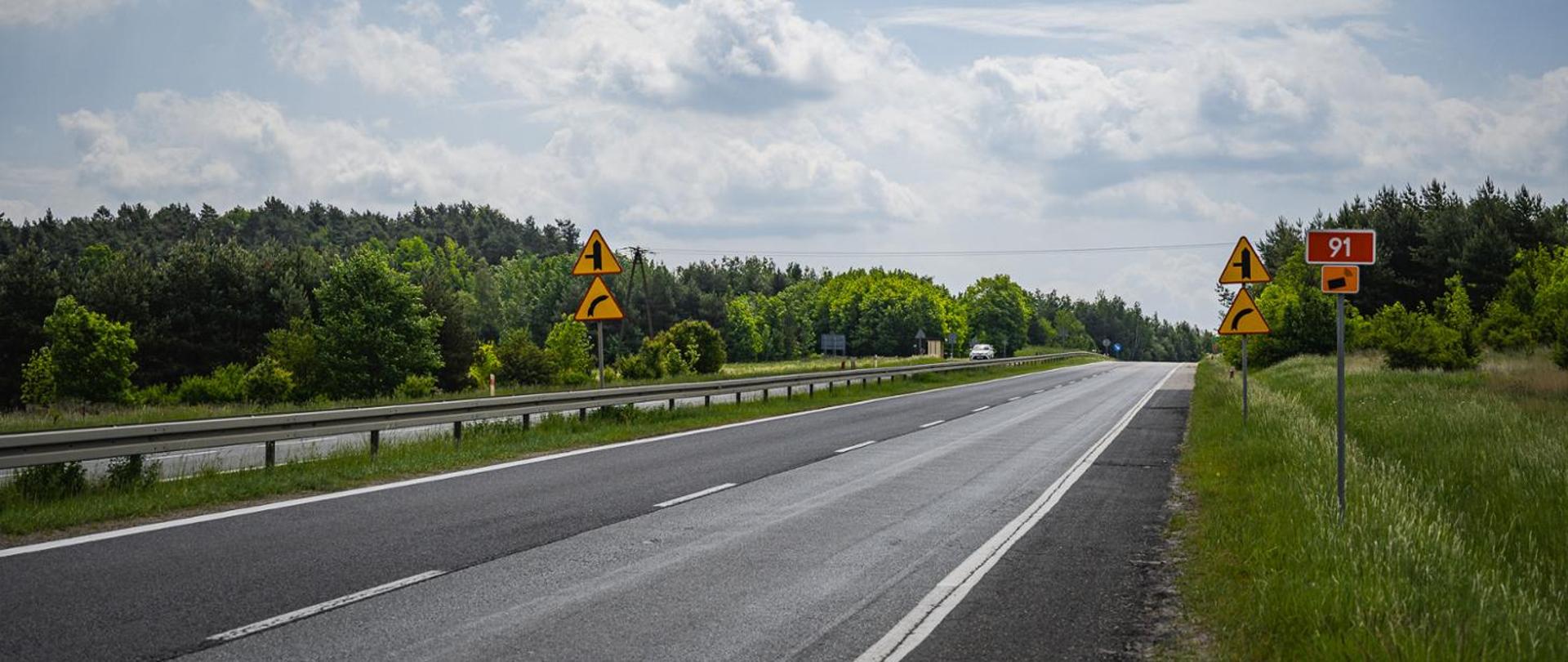 DK91 biegnąca przez teren otoczony zielenią i lasem. Jezdnie oddzielone są barierą energochłonną, a asfaltowa nawierzchnia prowadzi lekko pod górę. Po prawej stronie drogi rośnie wysoka trawa i znajdują się znaki drogowe, w tym czerwony znak z numerem drogi „91” oraz znaki ostrzegawcze informujące o zakręcie w prawo i wlocie drogi podporządkowanej (od strony lewej). W oddali na drodze porusza się samochód, a nad całością rozciąga się niebo z rozproszonymi chmurami.