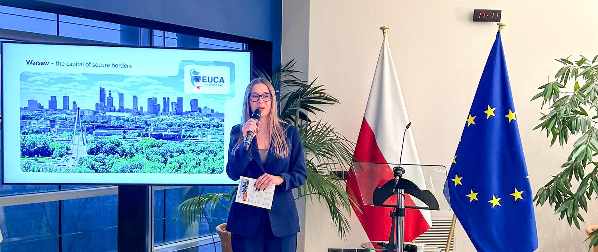 Osoba w garniturze prezentuje slajd o Warszawie na dużym ekranie, stojąc obok flag Polski i Unii Europejskiej.
