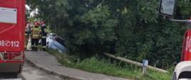 Ustabilizowany samochód osobowy fiat nad zboczem kanału w zaroślach. Trzech strażaków znajdujących się obok pojazdu. W tle kierowcy pojazdów oraz 2 samochody gaśnicze.