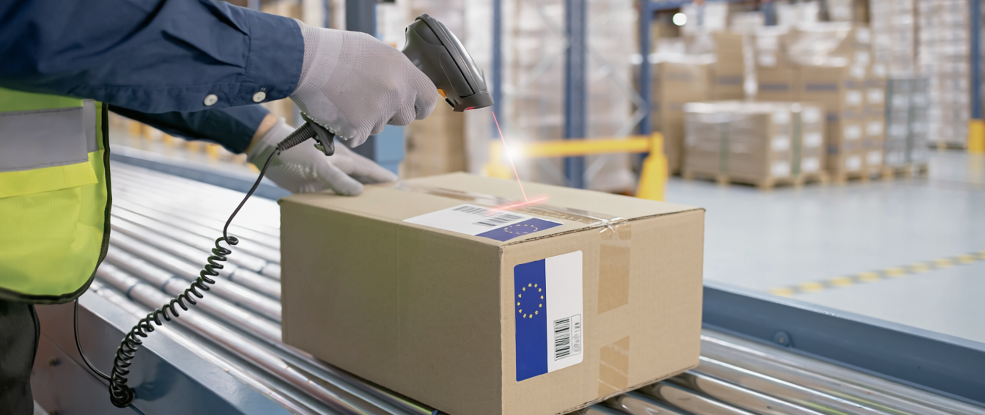 Osoba w kamizelce odblaskowej skanuje kod kreskowy na kartonie, leżącym na taśmie transportowej w magazynie pełnym regałów z zapakowanymi przesyłkami.