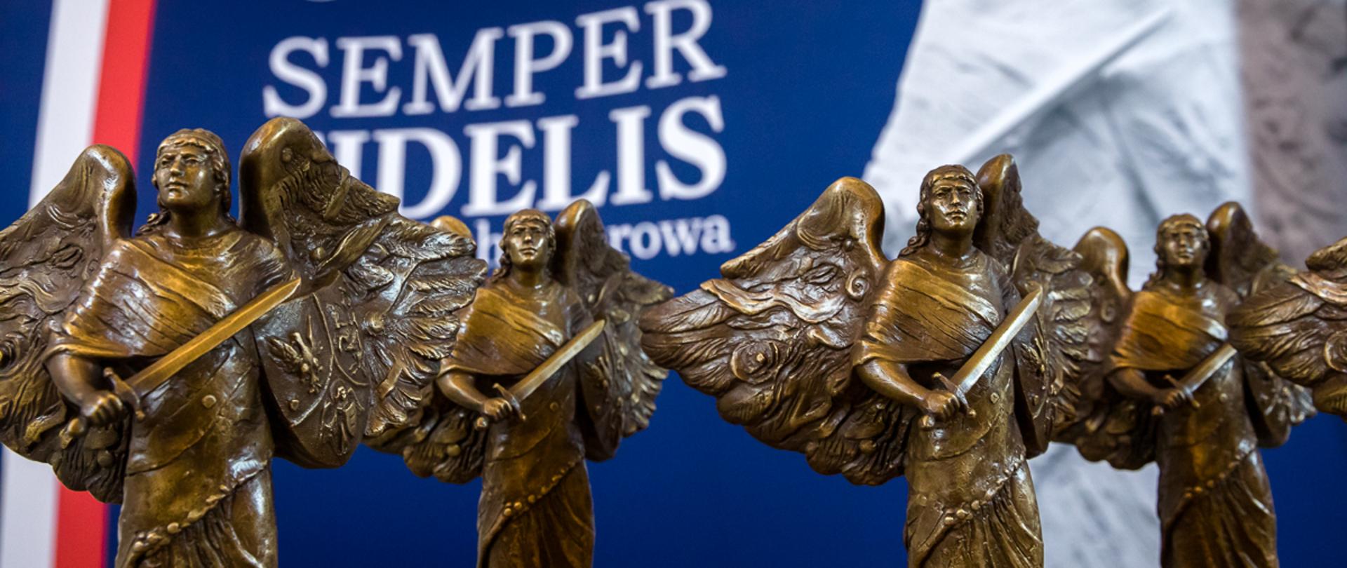 Cztery statuetki przedstawiające anioły z tarczą i mieczami. W tle niewyraźny napis Semper Fidelis.