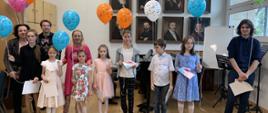 Uczniowie i nauczyciele z dyplomami i ozdobnymi balonami stoją na tle portretów wielkich kompozytorów.