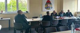 Sala konferencyjna podczas spotkania służbowego strażaków. Kilku mężczyzn w mundurach siedzi przy ustawionych w podkowę stołach, prowadząc naradę. Na ścianie widoczne jest godło Państwowej Straży Pożarnej, a w pomieszczeniu znajdują się okna, projektor sufitowy oraz butelki z wodą i dokumenty na stołach.