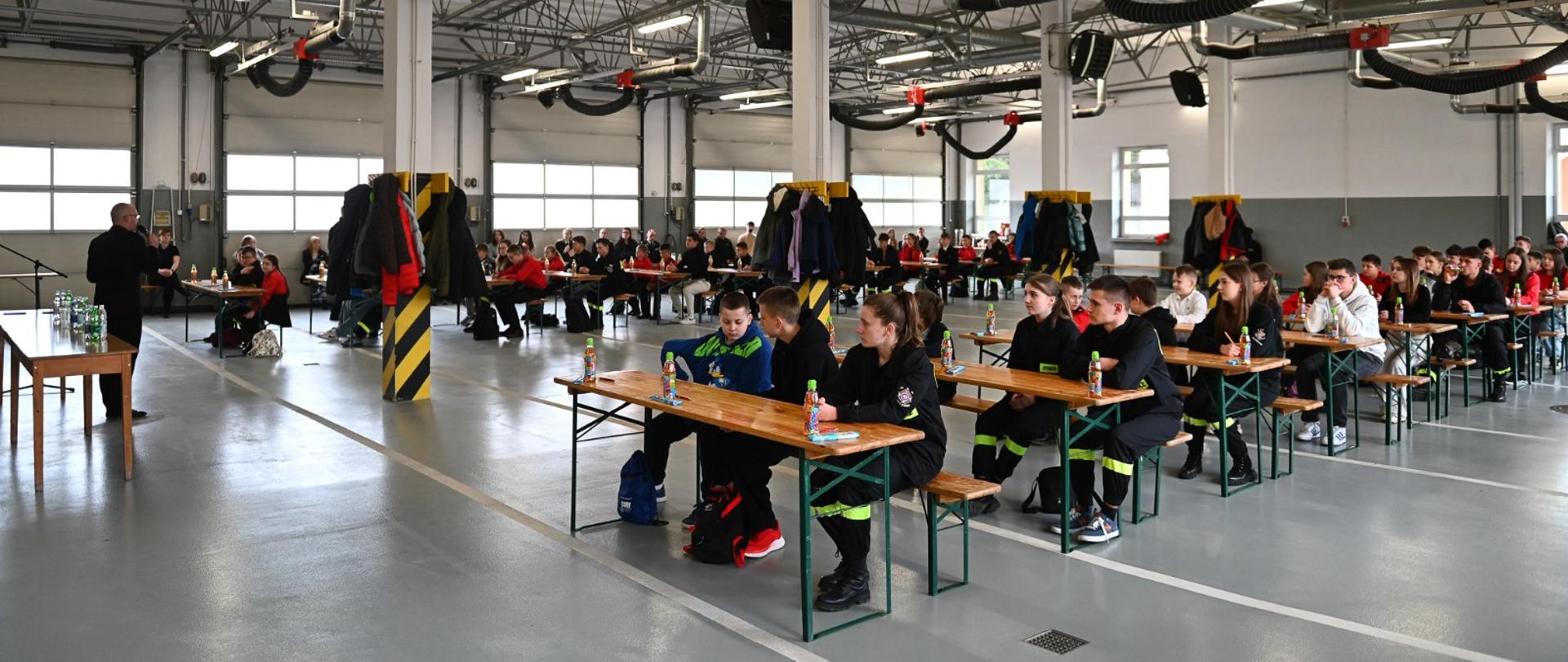 W ławkach siedzą uczestnicy konkursu, ławki ustawione są na garażu Jednostki Ratowniczo Gaśniczej. W garażu jest szara podłoga w tle widać bramy garażowe.