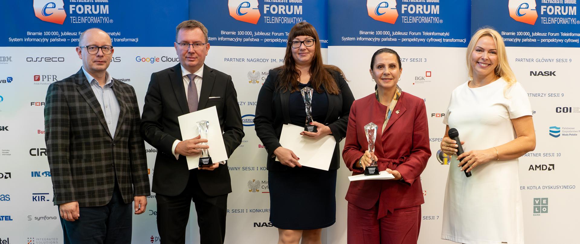 Trzy kobiety i dwóch mężczyzn stoją na tle ścianki Forum Informatyki. Stojący w środku mężczyzna i dwie kobiety trzymają w rękach dyplomy i kryształowe statuetki. Wszyscy uśmiechają się do zdjęcia.