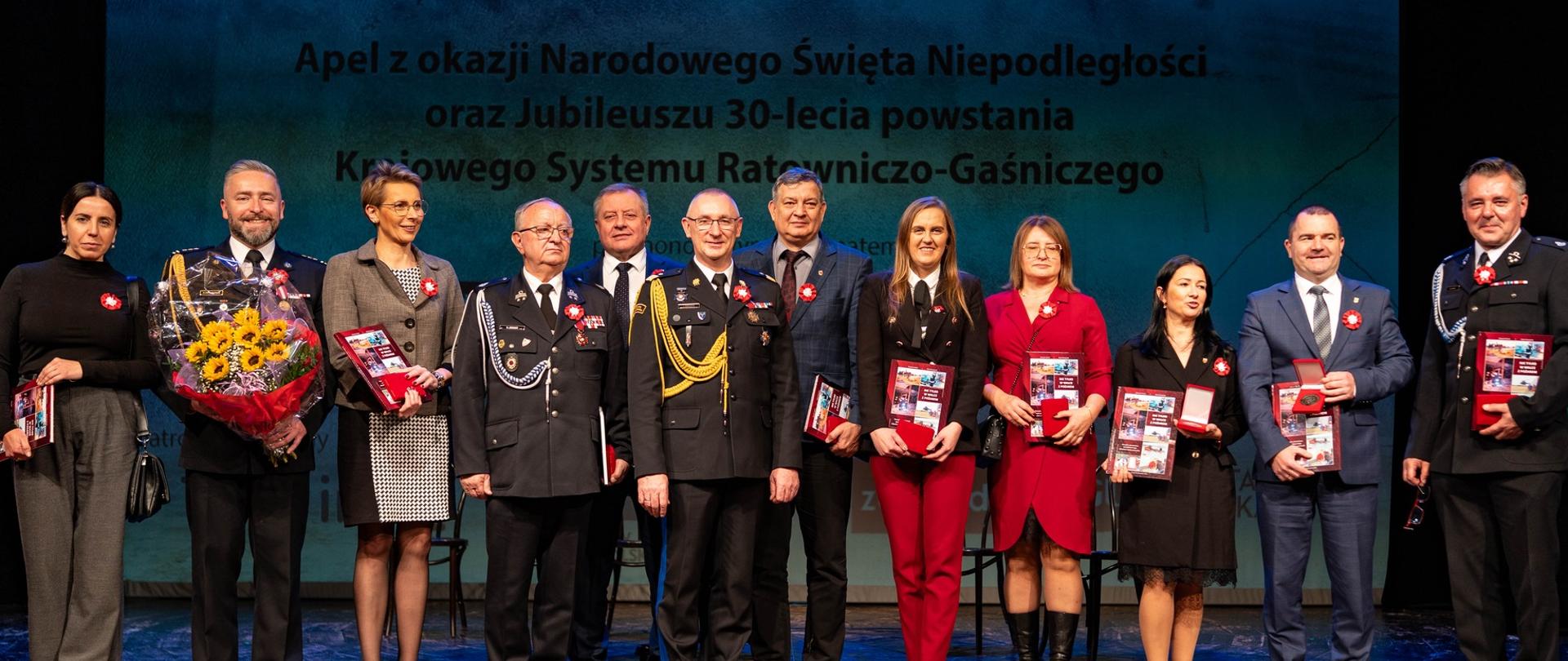 osoby wyróżnione medalem jubileuszowym 30-lecia KSRG ustawione w szeregu na scenie teatru prezentujące w dłoniach medale i książki