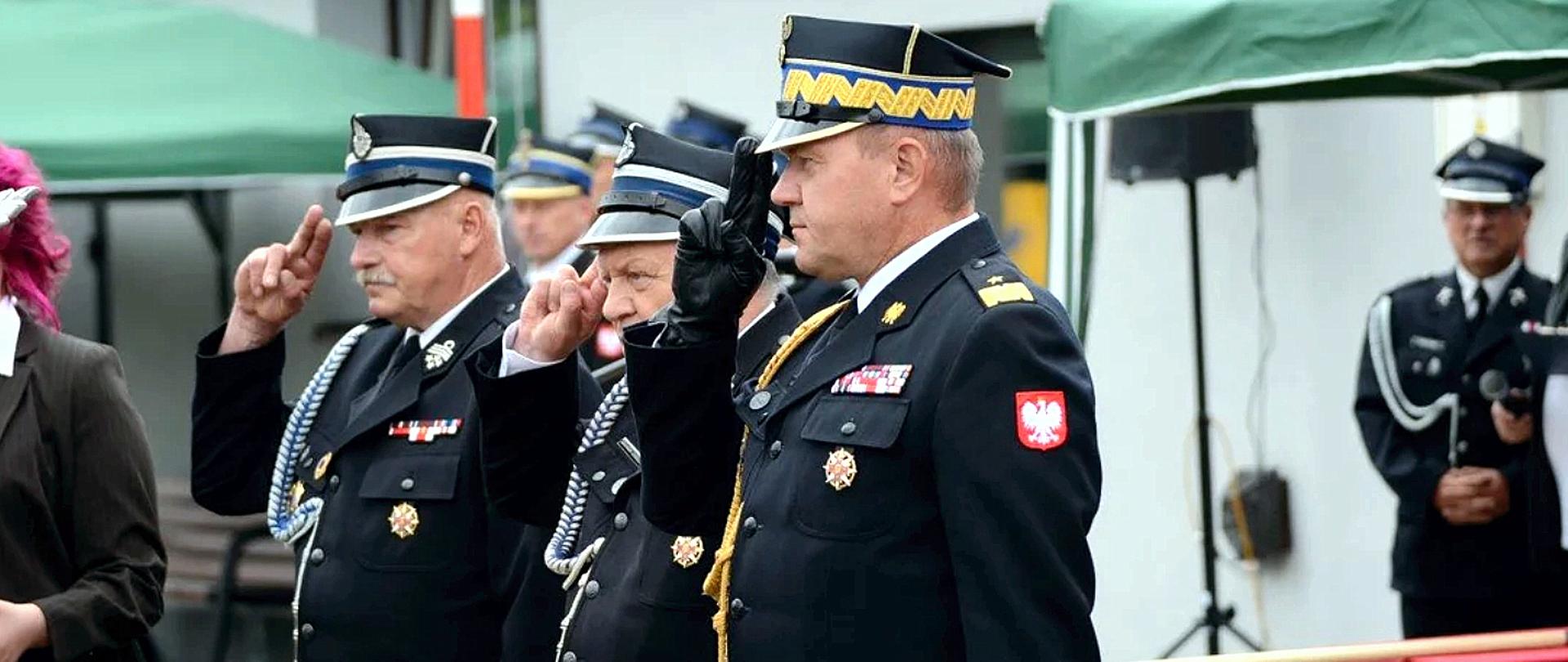 Zdjęcie zrobione na zewnątrz, podczas uroczystości. W szeregu stoi 3 strażaków, dwóch z nich to strażacy ochotnicy. Trzeci, to oficer Państwowej Straży Pożarnej w stopniu generała. Strażacy ubrani są w mundury wyjściowe ze sznurami galowymi. Strażacy salutują.