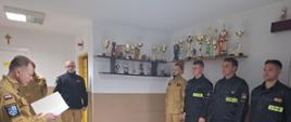 Starszy brygadier Maciej Suchomski (po lewej) trzyma w rękach pismo rozkazu służbowego, młodszy brygadier Dariusz Chylewski ubrani w mundury służbowe oraz strażacy ze zmiany służbowej w mundurach koszarowych w szeregu pod ścianą zgromadzeni w pomieszczeniu świetlicy Komendy Powiatowej Państwowej Straży Pożarnej w Sępólnie Krajeńskim. 