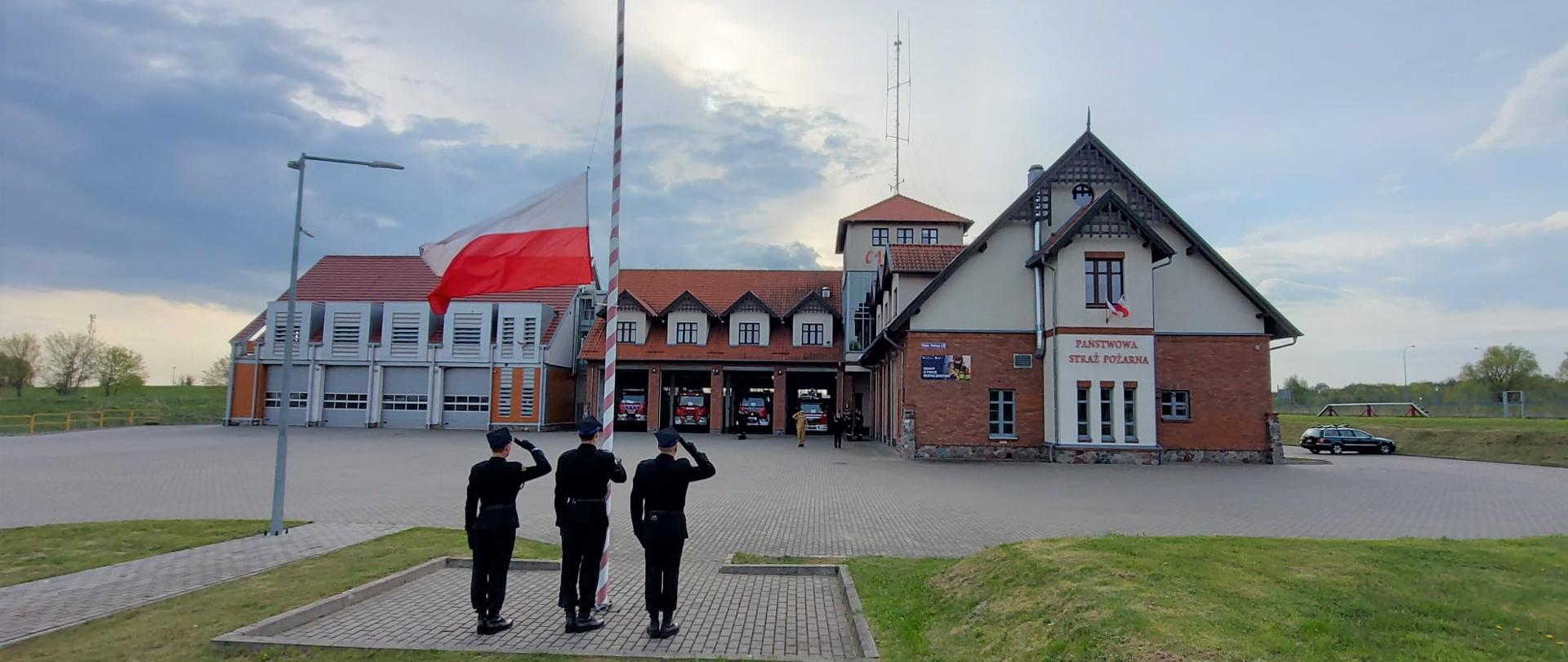 Zdjęcie przedstawia strażaków Komendy Powiatowej Państwowej Straży Pożarnej w Gołdapi podczas podnoszenia flagi Rzeczypospolitej Polskiej przy akompaniamencie hymnu państwowego z okazji Dnia Flagi Państwowej Rzeczypospolitej Polskiej. Dowódcy zmian, funkcjonariusze nie będący w pododdziale oraz dowódca pocztu flagowego i asystujący oddają honory przez salutowanie. W tle widoczny jest również obiekt strażnicy i pojazdy pożarnicze