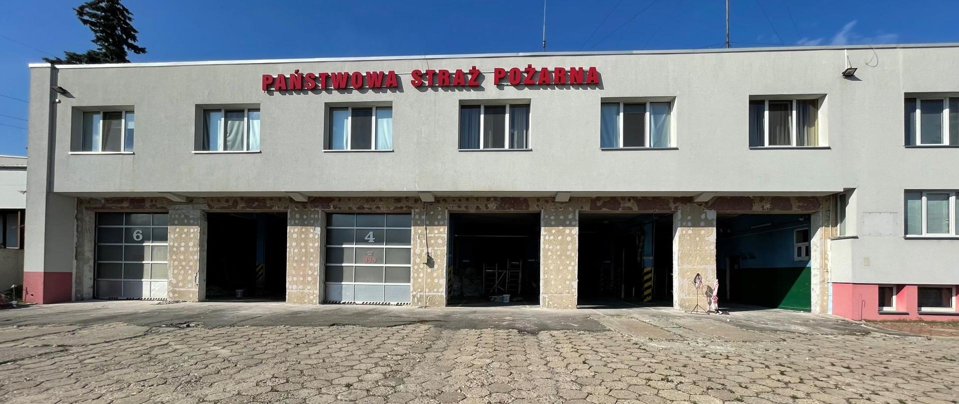 Plac z kostki. W oddali budynek garażowy z częściowo usuniętymi bramami wjazdowymi. Na budynku widnieje napis Państwowa Straż Pożarna