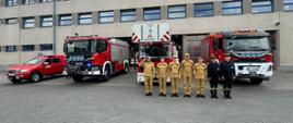 Strażacy ze zmiany JRG ustawieni w szeregu przed bramami garażowymi jednostki, za nimi widoczne samochody bojowe z włączonymi sygnałami świetlnymi