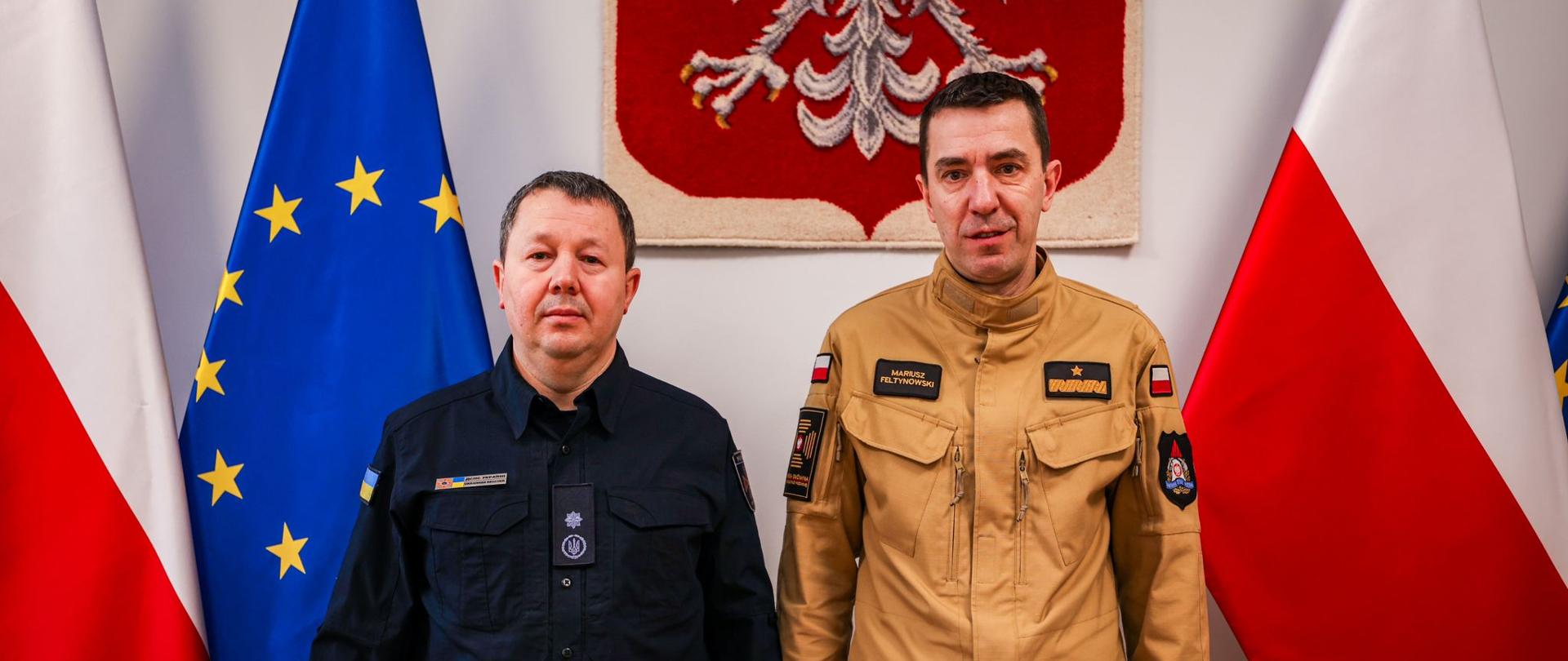 Dwóch mężczyzn w mundurach, jeden w granatowym mundurze, drugi w piaskowym mundurze pozują do zdjęcia. W tle godło Polski, z prawej, biało-czerwona flaga Polski, z lewej, biało-czerwona flaga Polski i niebieska flaga Unii Europejskiej.