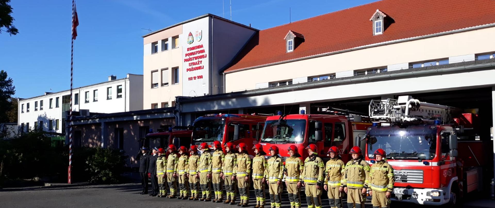 ODDALI HOŁD ZMARŁEMU STRAŻAKOWI. Na zdjęciu widać strażaków przed budynkiem Komendy, samochody strażackie wyjechały z garażu, mają włączone sygnały świetlne i dźwiękowe, widać maszt z flagą Polski i budynek Komendy Powiatowej PSP w Kościanie