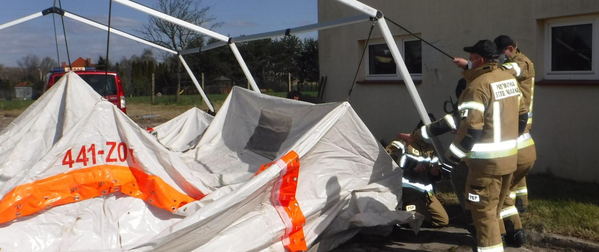Na zdjęciu widać trzech strażaków w żółtych strojach i czarnych czapkach z daszkiem rozkładających namiot. W tle widać czerwony samochód strażacki, ścianę budynku oraz drzewa. Jest widno