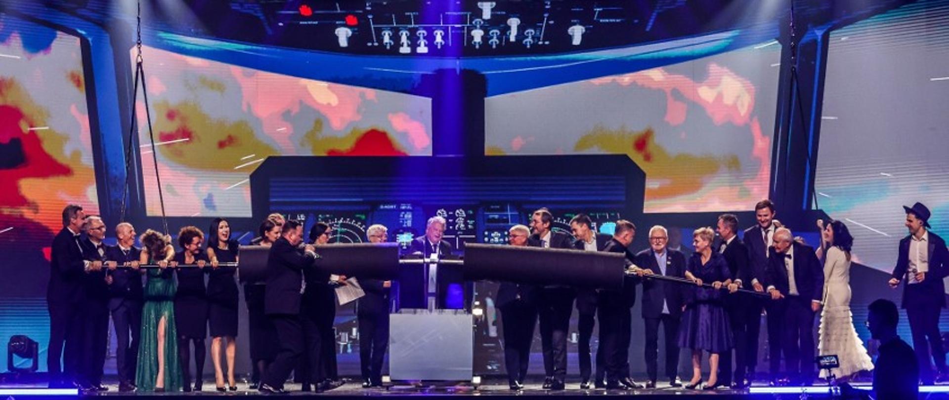 grupa osób stoi na scenie za nimi makieta kokpitu samolotu