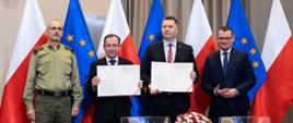 Przy białym stole stoi minister Czarnek, dwóch mężczyzn w garniturach i jeden w zielonym mundurze, minister Czarnek i mężczyzna w czarnym garniturze trzymają przed sobą dokumenty, przed nimi na stole mały bukiet biało-czerwonych kwiatów, za nimi rząd flag Polski i UE.