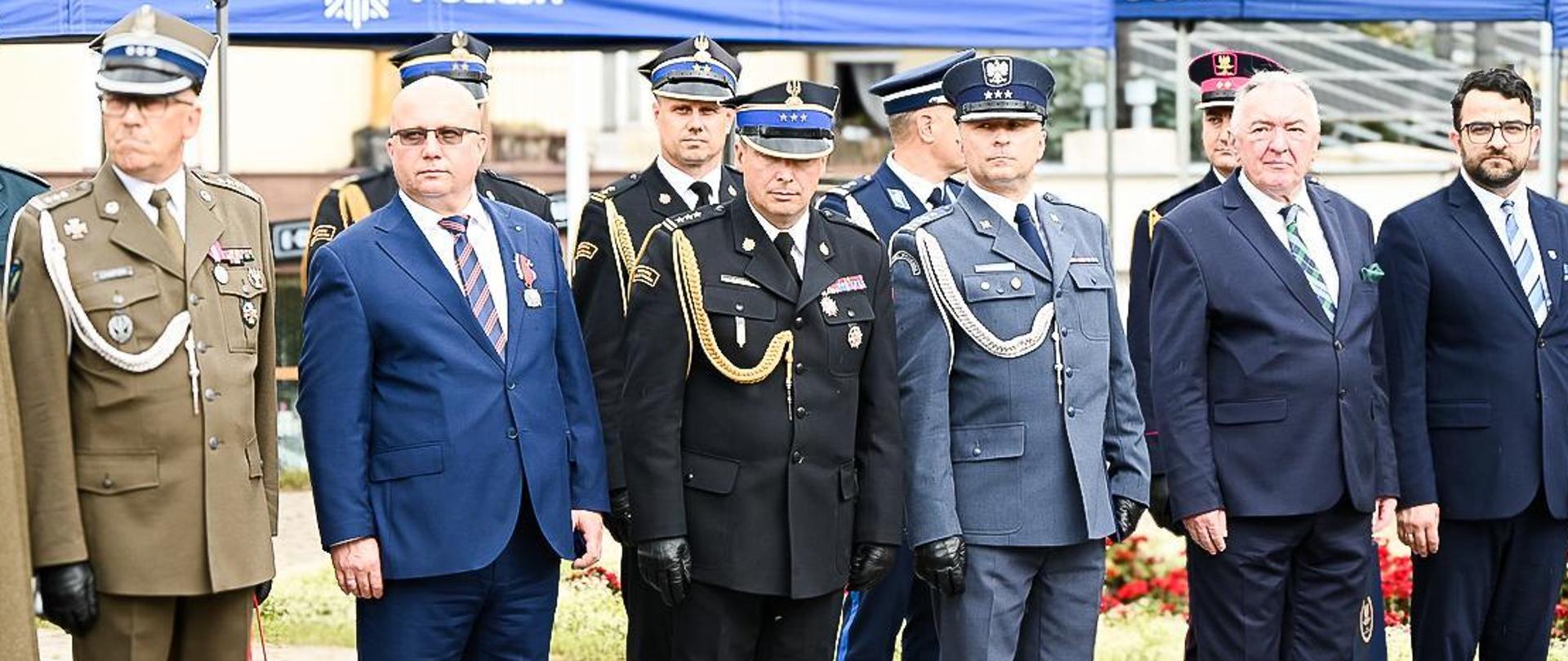 Fotografia wykonana podczas Wojewódzkich Obchodów Święta Policji w dniu 17 lipca 2024 roku na Plac Farny w Rzeszowie. Na zdjęciu stoją funkcjonariusze Policji, Państwowej Straży Pożarnej Żołnierze Wojska Polskiego oraz pozostali goście. Funkcjonariusze i żołnierze ubrani są w umundurowania galowe i czapki wyjściowe. W tle widać namiot koloru niebieskiego z napisem Policja. 